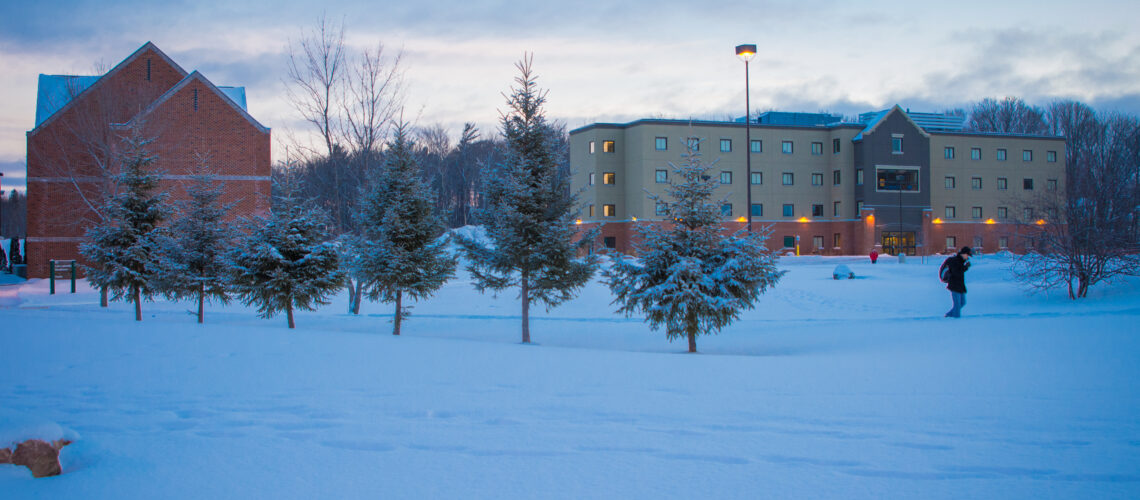 SSM Campus. Winter, evening. Snow covered residence building and person walking across campus through a path of snow lined with pine trees.