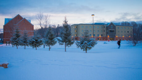 SSM Campus. Winter, evening. Snow covered residence building and person walking across campus through a path of snow lined with pine trees.